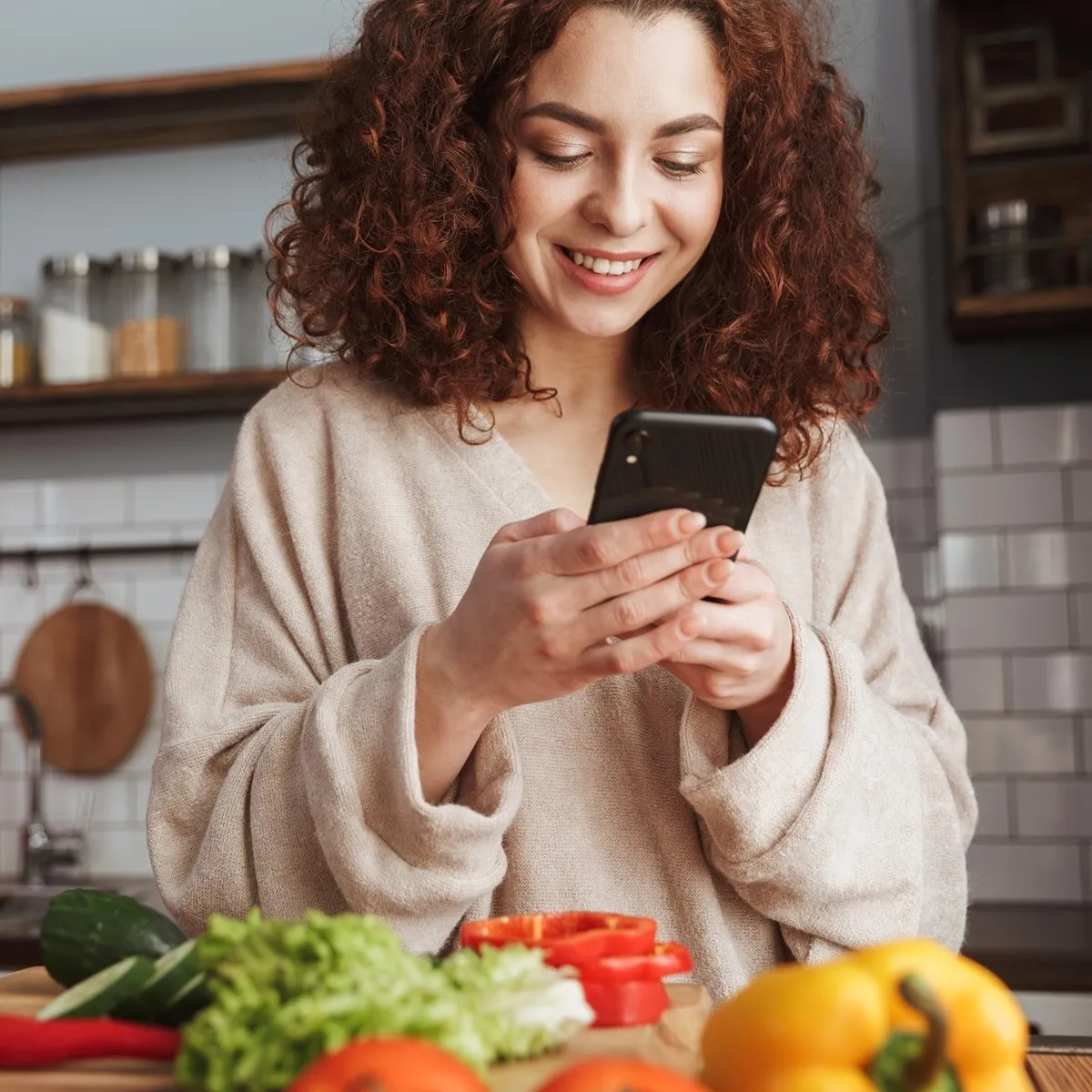 Young woman with curly hair smiling while taking a photo of fresh vegetables in a bright kitchen.