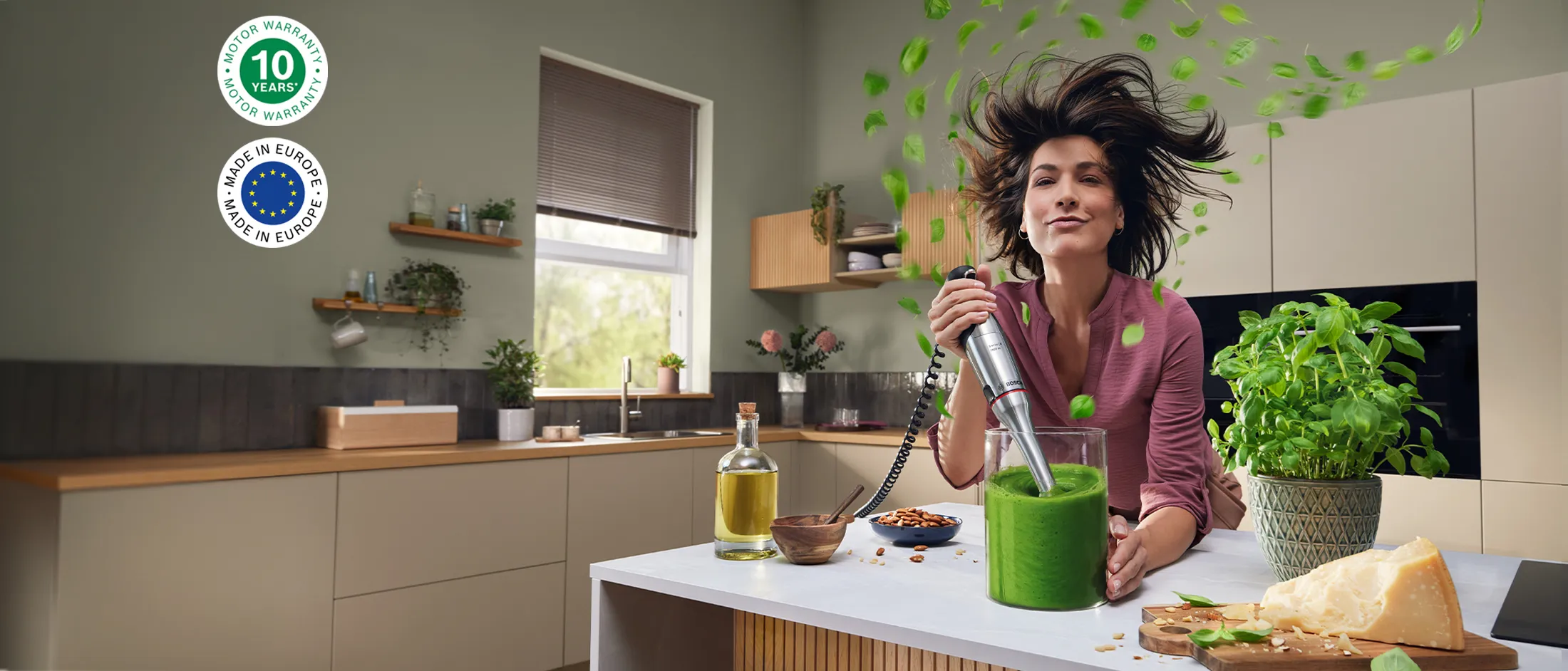 Una mujer sonriente prepara un batido verde en una cocina luminosa, con hojas frescas volando a su alrededor, rodeada de frutos secos, queso y albahaca.
