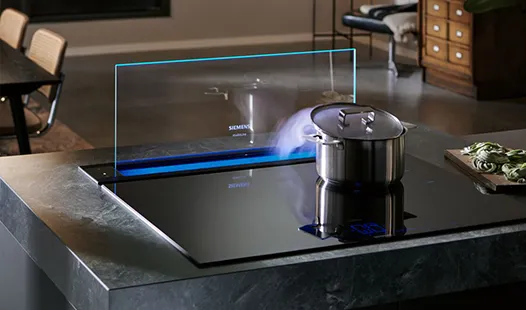 A black Siemens induction hob with a retractable glass downdraft extractor, which is lit with a blue light and is actively drawing steam from a stainless steel pot on the hob.