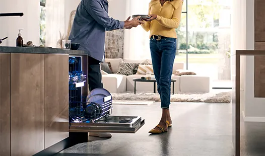 A man and woman stand together in a kitchen, next to an open dishwasher filled with clean dishes. 