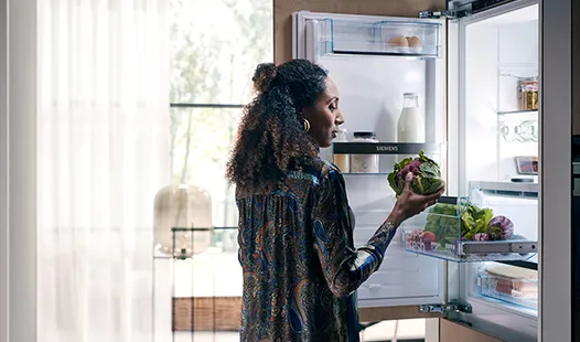A woman presents a colorful salad while standing in front of an open refrigerator stocked with groceries.  