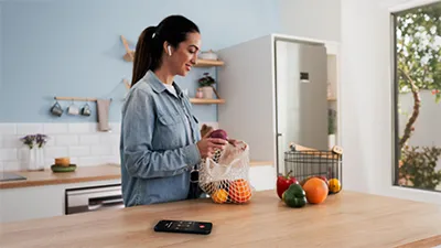 alt-A woman stands in a bright kitchen, holding a mesh bag of fruits, while contacting customer service on her smartphone placed on the table.