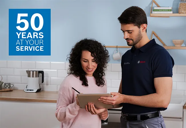 Een Bosch-technicus in handschoenen repareert een apparaat in een moderne keuken terwijl een vrouw aandachtig toekijkt. Gereedschap ligt nabij op het aanrecht.