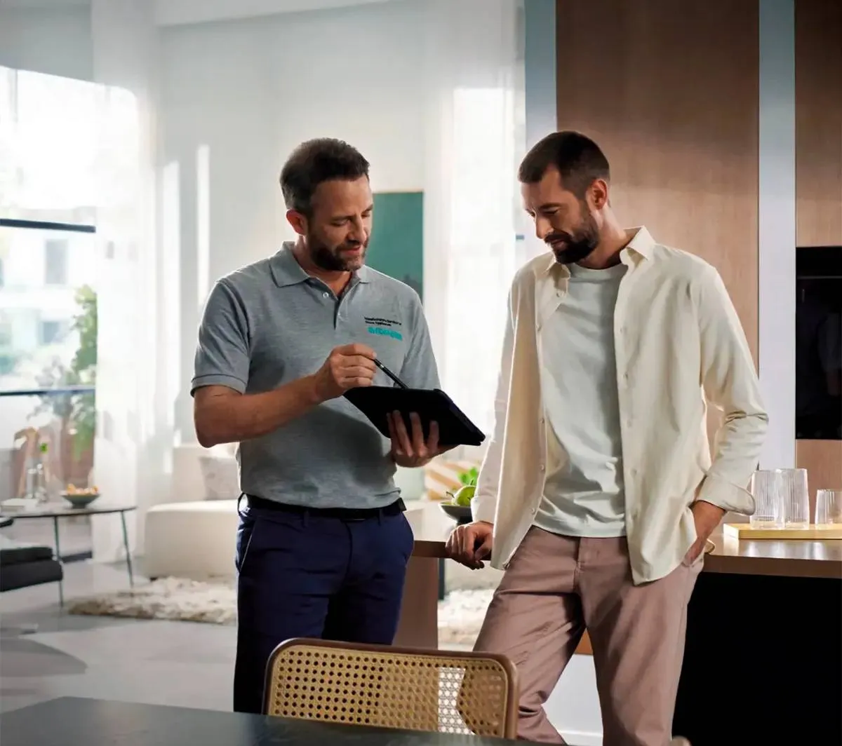 Deux hommes discutent, l'un tenant une tablette à la main, dans un salon moderne et lumineux avec des meubles élégants.