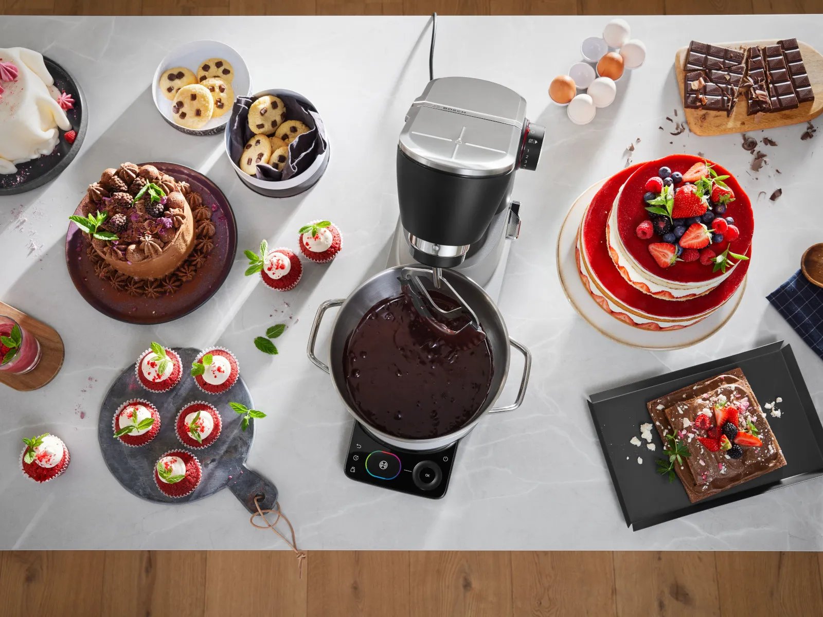 Vue plongeante d’un robot pâtissier fouettant du chocolat sur un plan de travail en marbre, entouré d’un assortiment de gâteaux, cupcakes et biscuits.