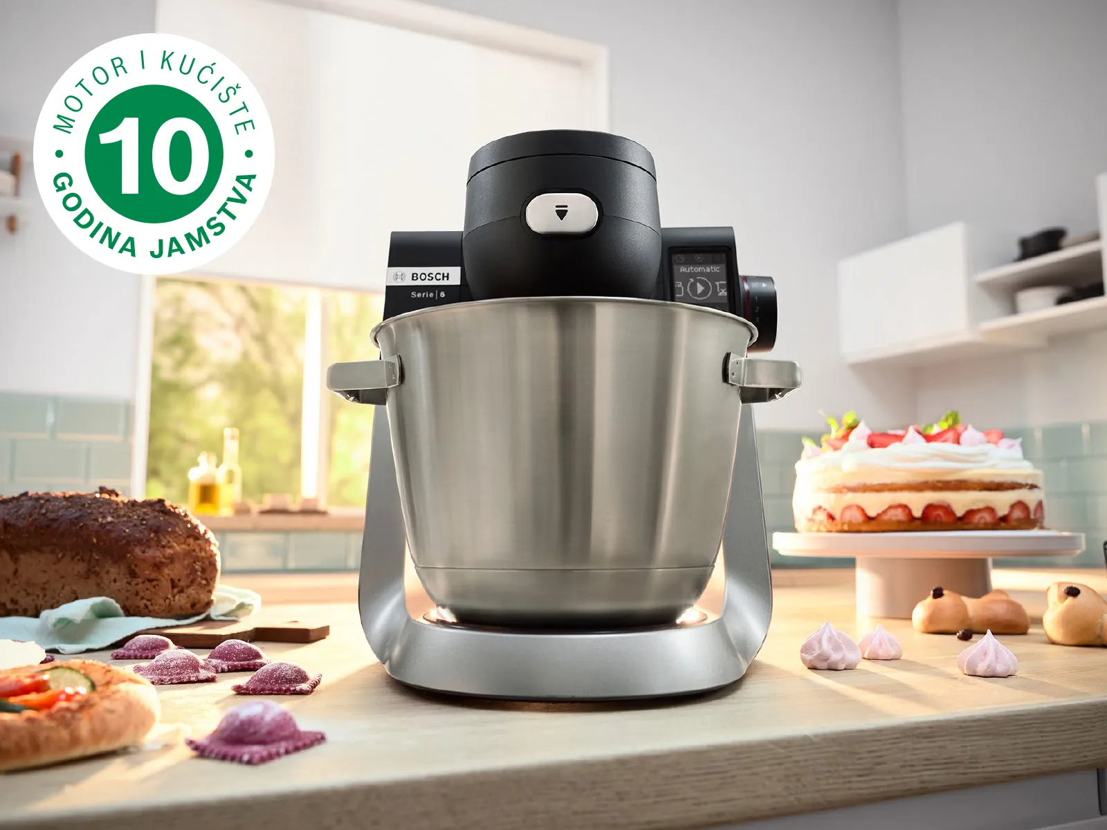 Slika prikazuje samostojeći mikser serije 6 na kuhinjskoj radnoj površini pokraj kojega se nalazi nekoliko kugli tijesta i umetnuti logotip 10-godišnjeg jamstva.