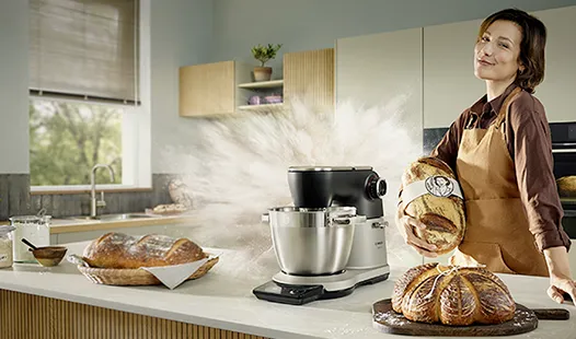 Frau in der Küche mit Bosch Mixer und frisch gebackenem Brot auf der Arbeitsplatte, umgeben von Zutaten.