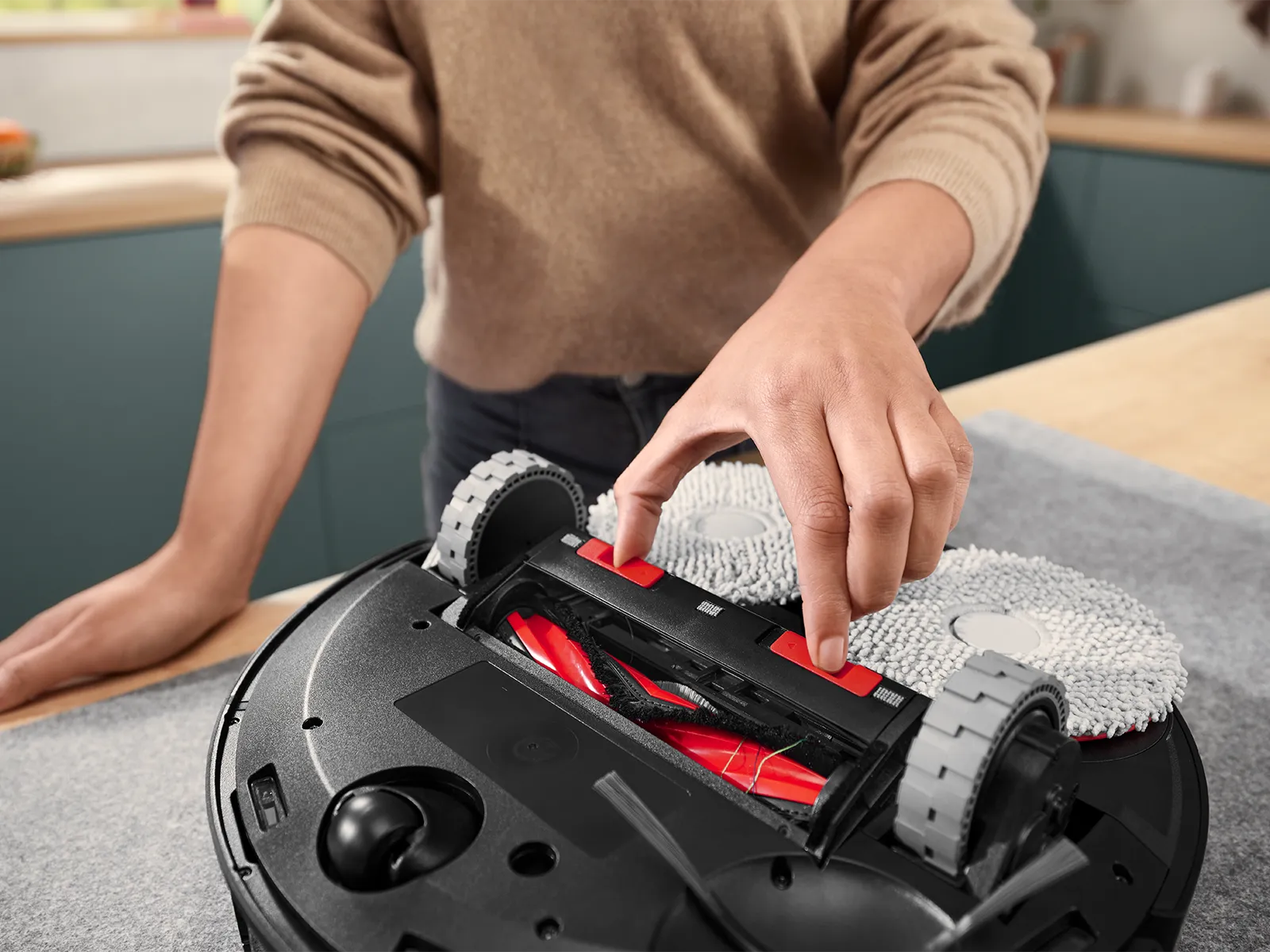 A person adjusts the brush roll of an upside-down robotic vacuum on a countertop.