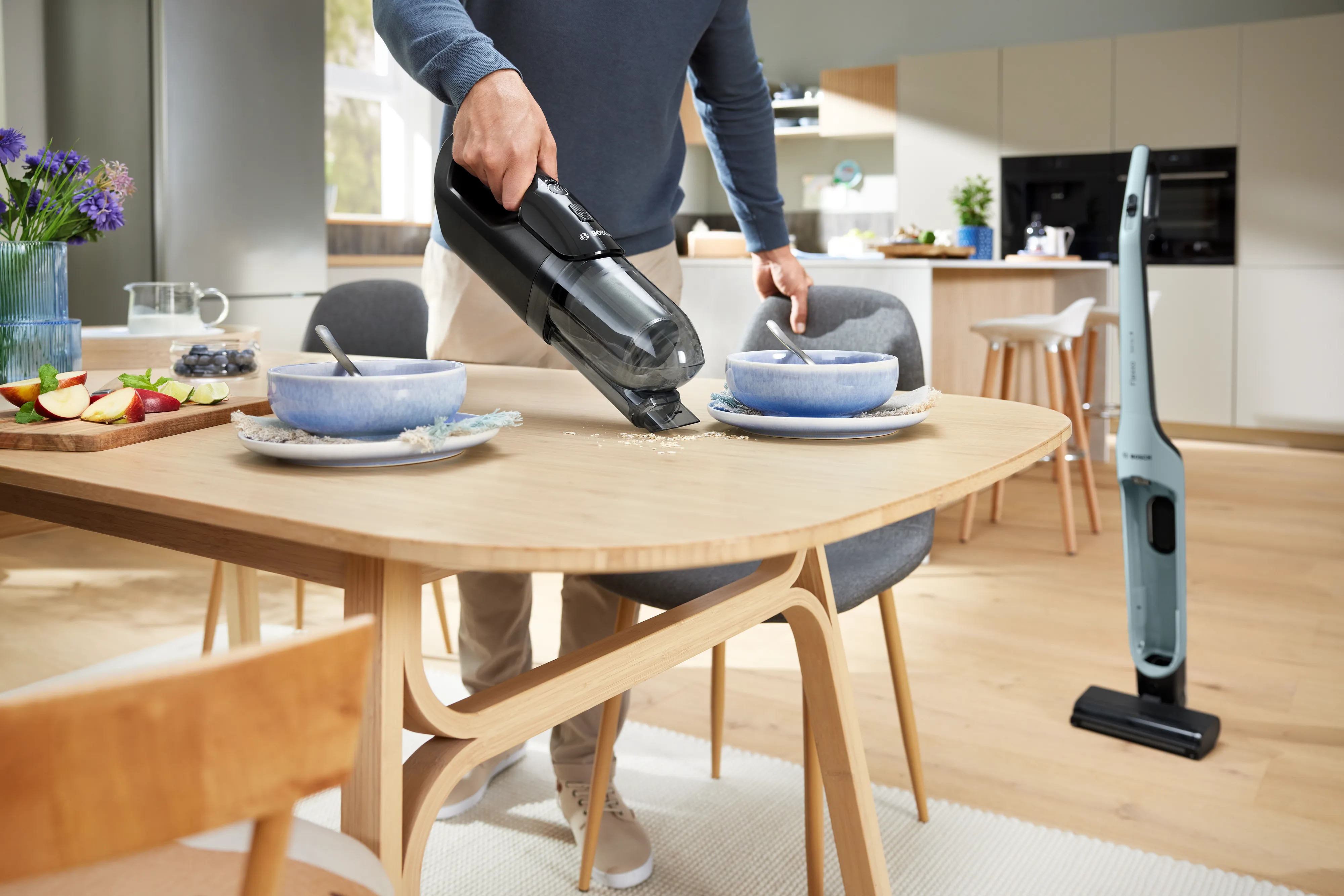 Eine Person saugt mit dem Handstaubsauger Krümel von einem Holztisch auf. Im Hintergrund steht der Flexxo Serie 4 Akku-Staubsauger in einer hellen Küche.