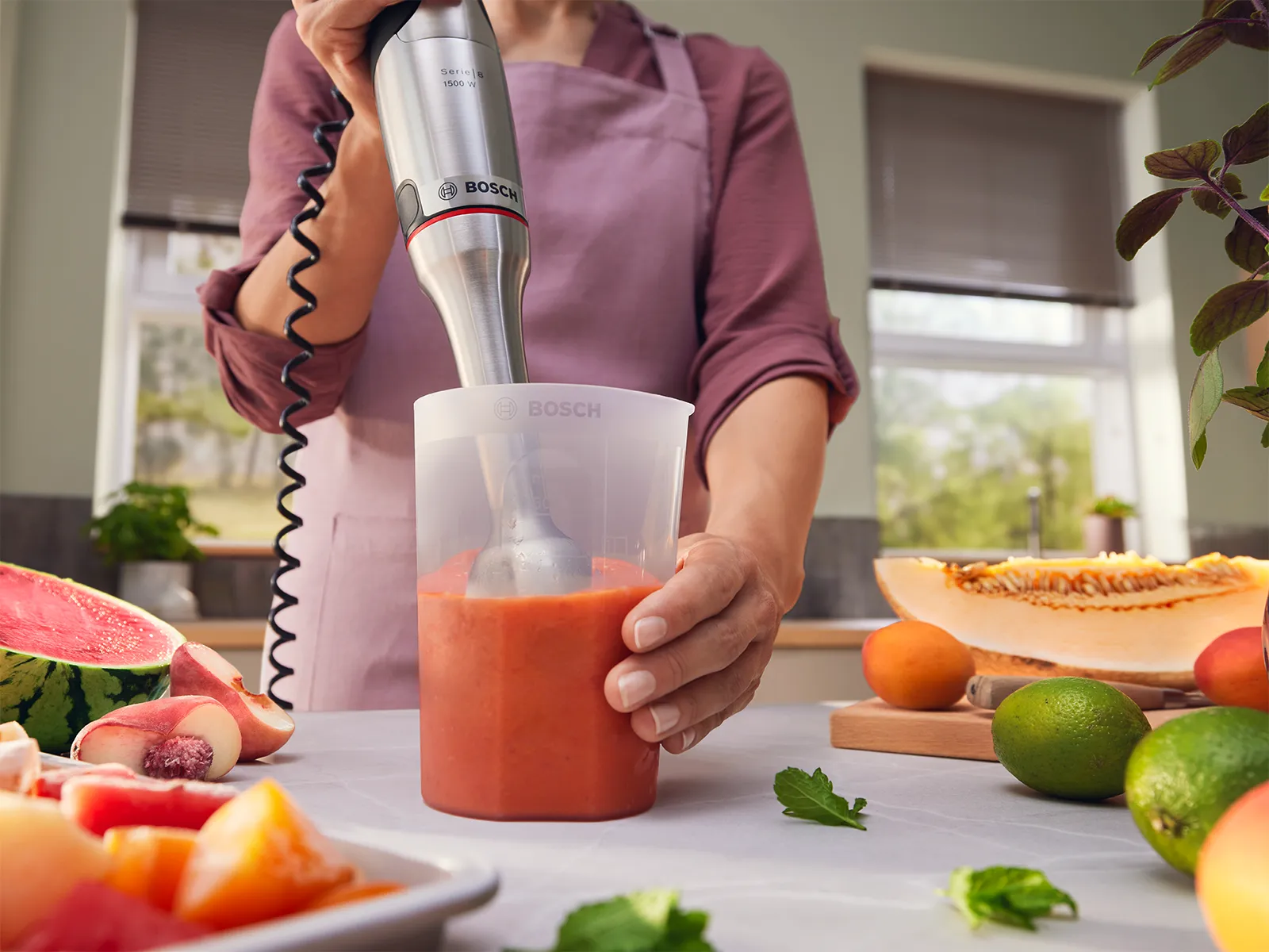 erson in a light purple apron using a Bosch hand blender to blend chopped fruit in a clear beaker, with assorted fresh fruits on a cutting board in a bright kitchen.