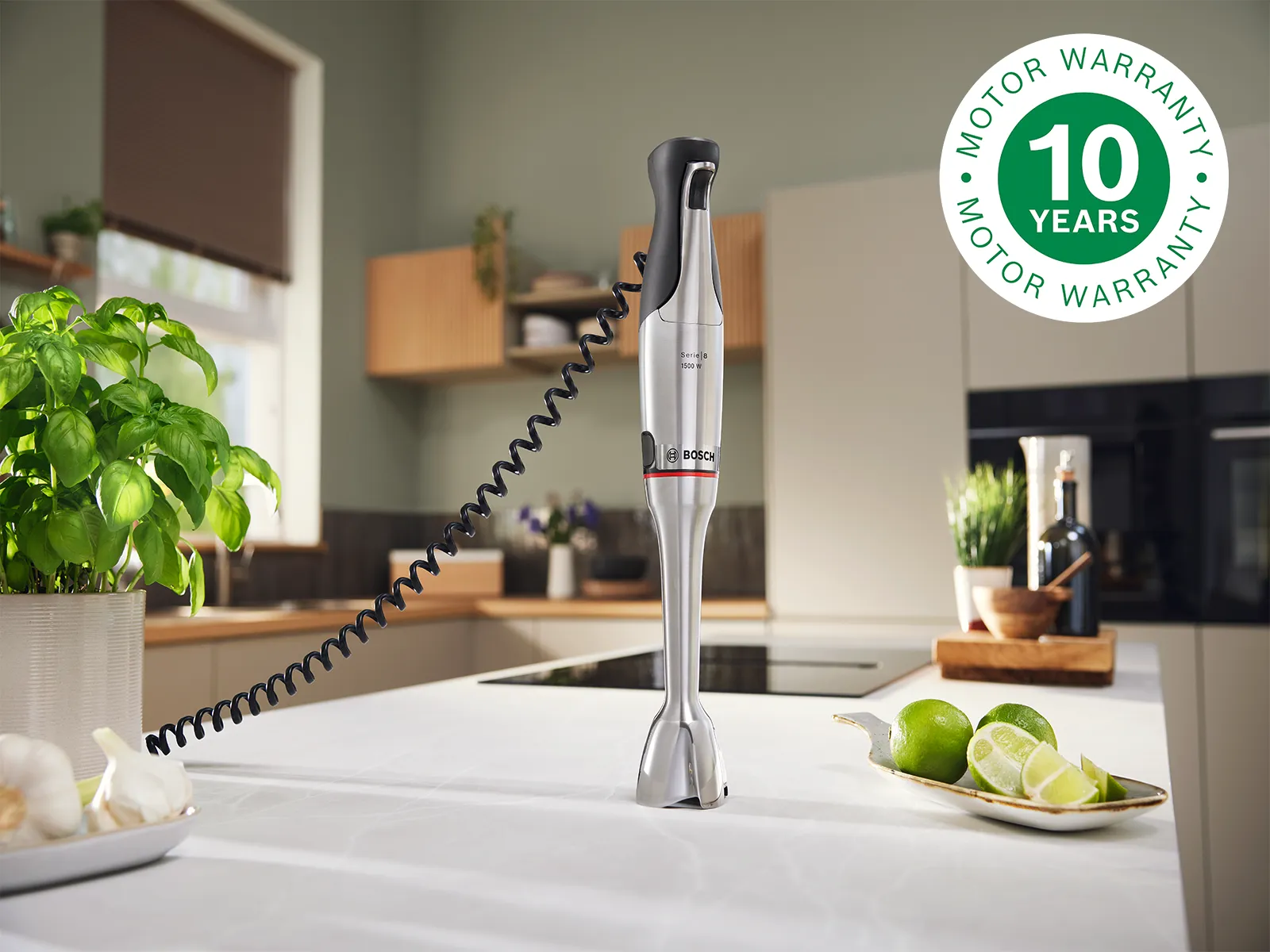 Licuadora de mano sobre la encimera de la cocina con planta de albahaca, dientes de ajo y rodajas de lima, en un entorno de cocina limpio y con el logotipo de garantía del motor de 10 años.
