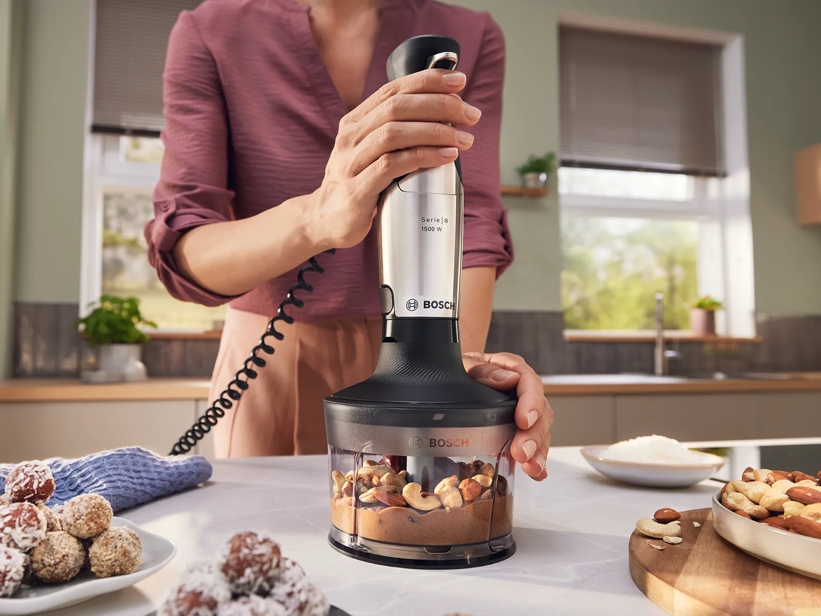 Bir şəxs tərəvəzləri doğramaq üçün doğrayıcı aksesuardan istifadə edərək Bosch əl blenderini tutub.