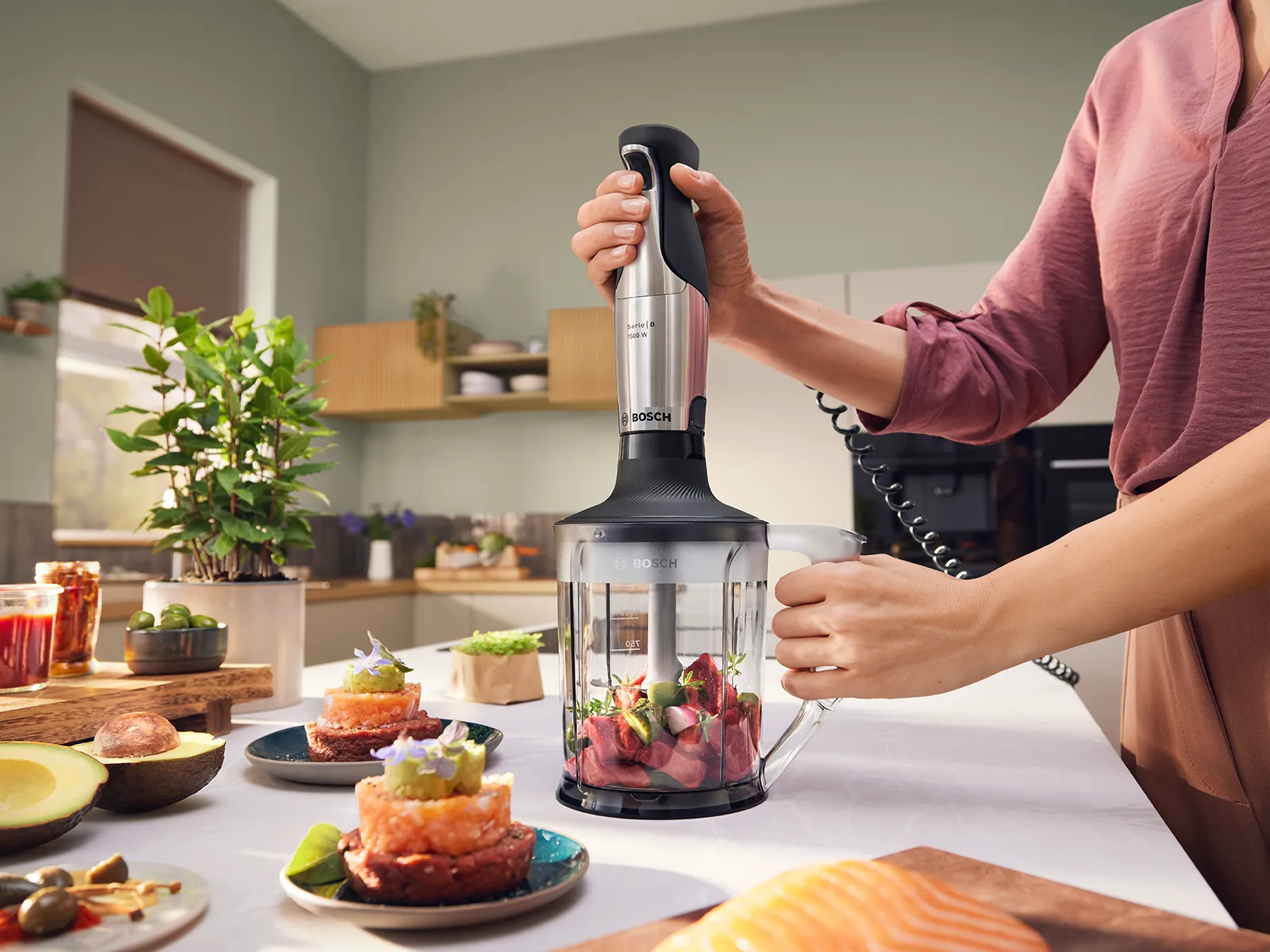 Donna con camicetta viola chiaro che usa un frullatore a immersione Bosch su un contenitore trasparente per preparare una tartare di manzo alle erbe aromatiche; antipasti, olive e avocado sono sistemati su un elegante piano di lavoro in una cucina luminosa.