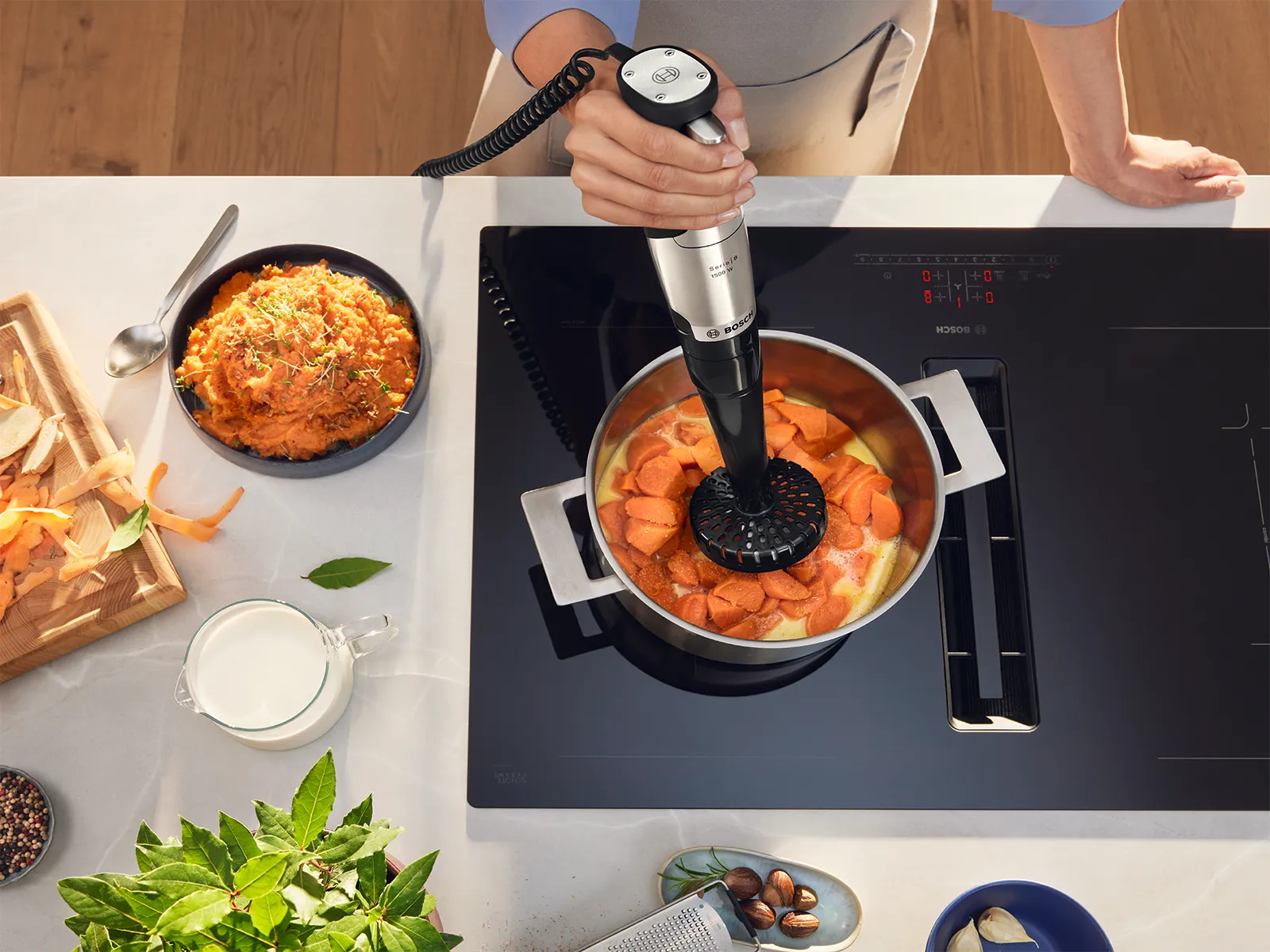 Persona che utilizza un frullatore a mano metallico con attacco schiacciatore in una pentola piena di miscela di arance su piano cottura elettrico, con bucce di verdure, zenzero, erbe aromatiche, latte e spezie su un luminoso bancone da cucina moderno.