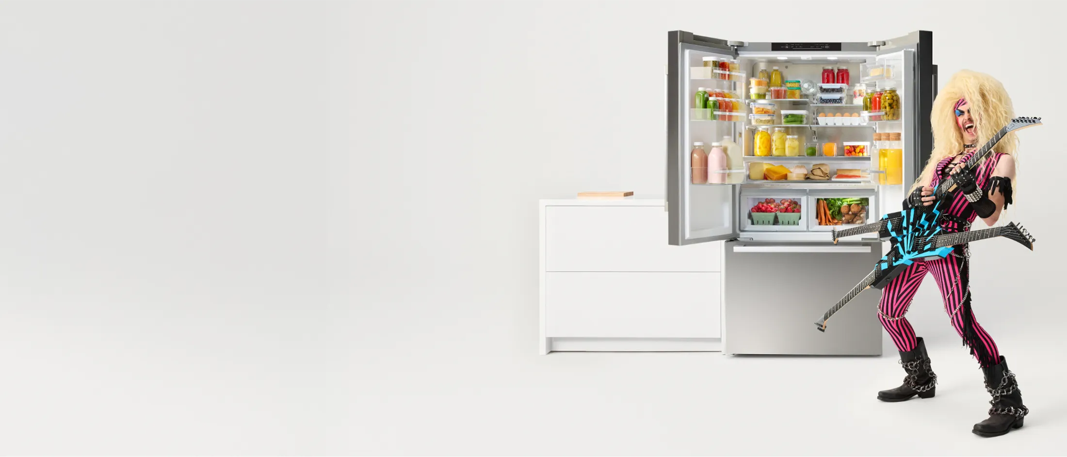 A rockstar with big hair and a guitar stands beside a Bosch open refrigerator filled with colorful food and drinks. A modern white dresser is nearby.