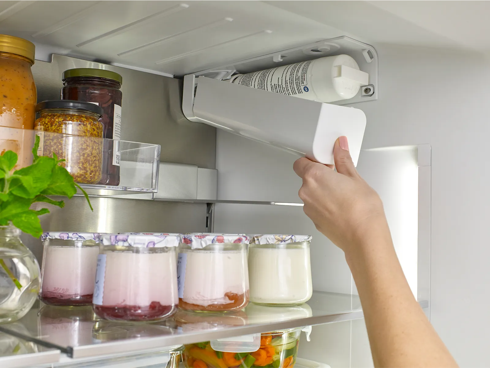 Hand replacing a water filter in a Bosch refrigerator, next to jars and yogurt containers.