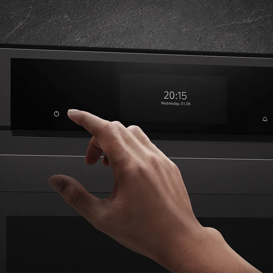 Primo piano di una mano che preme il pulsante di accensione sul pannello di controllo di un forno digitale che mostra data e ora.