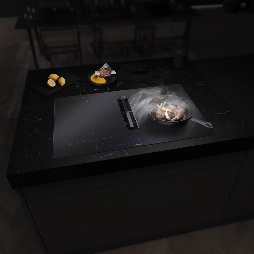 Black kitchen island with a modern induction cooktop, frying meat in a skillet, and dishes with lemons and sauce in the background.
