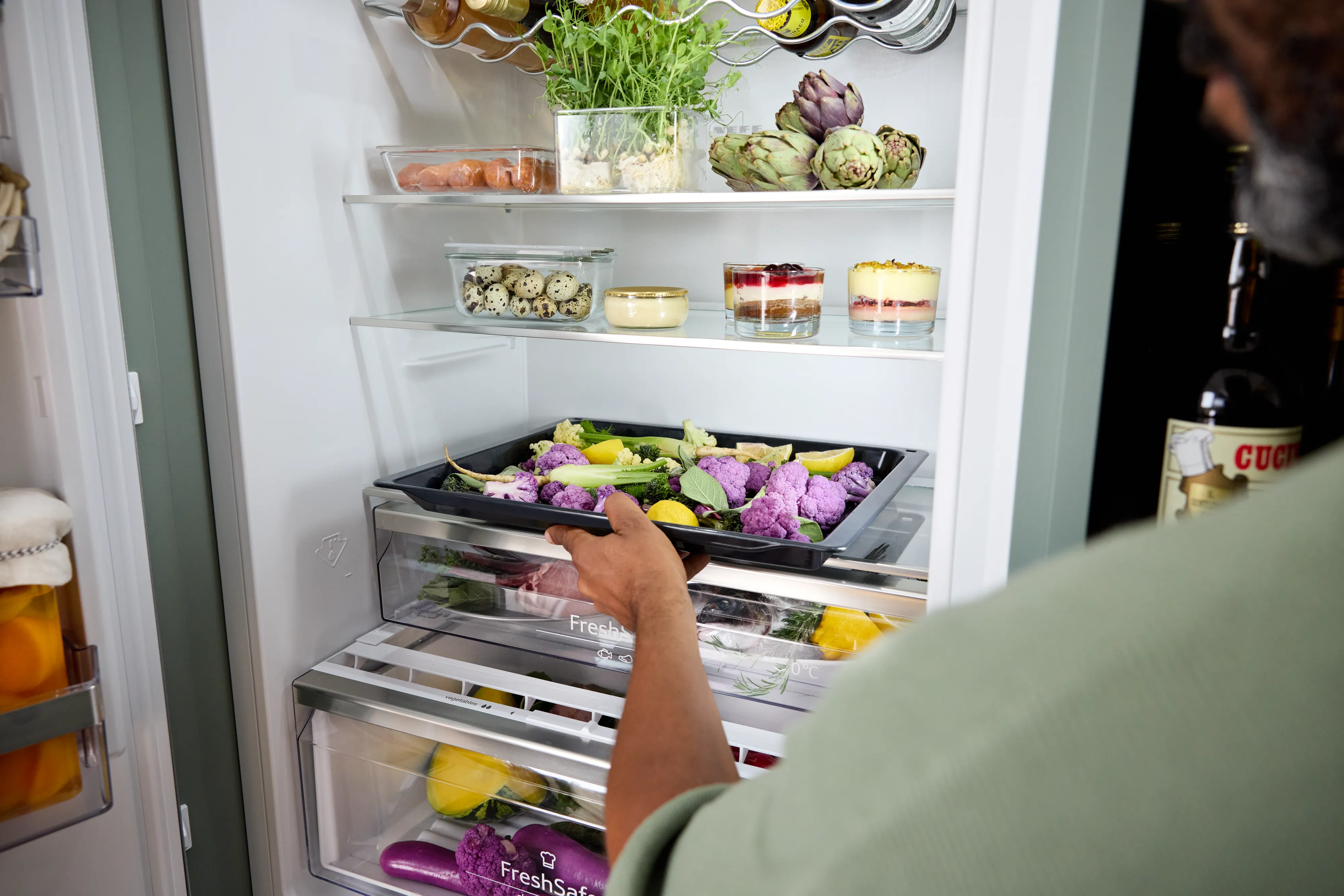 Person schiebt ein Backblech mit lila Blumenkohl und anderem Gemüse in eine Kühlschrankschublade. Im Innenraum sind oben frische Kräuter, Artischocken, Glasbehälter mit Wachteleiern und Dessertgläser zu sehen; an der Tür befinden sich Flaschen.