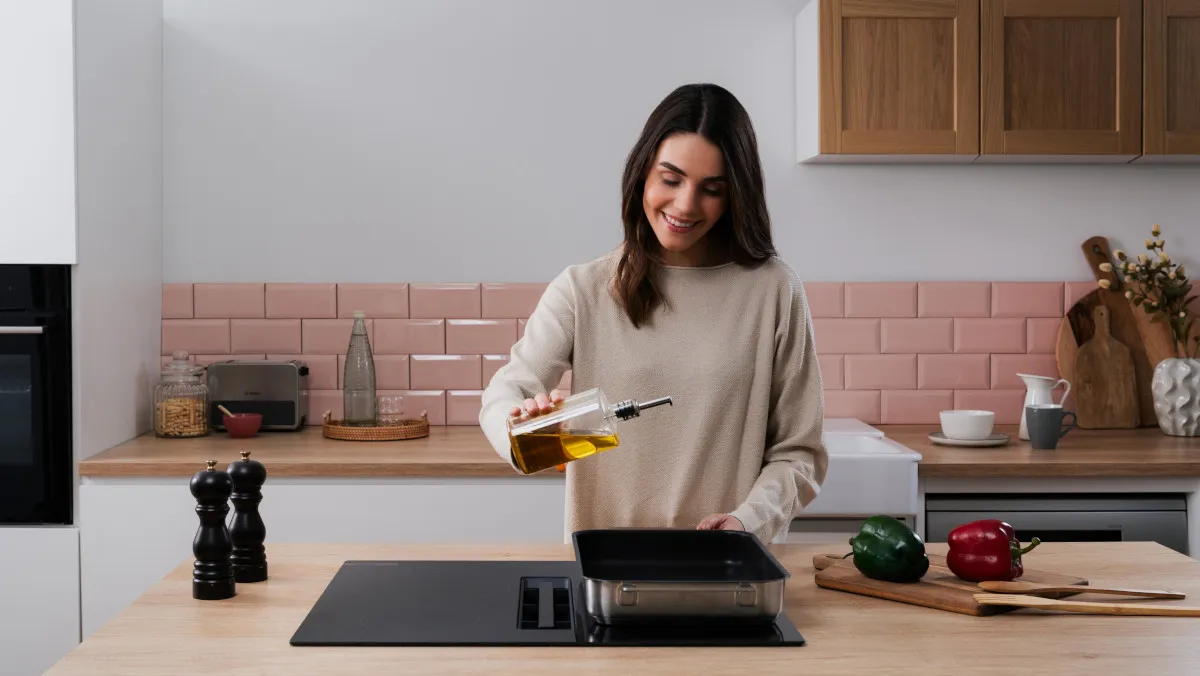 Femme souriante versant de l'huile dans une poêle sur une plaque de cuisson dans une cuisine moderne avec plan de travail en bois et carrelage rose.