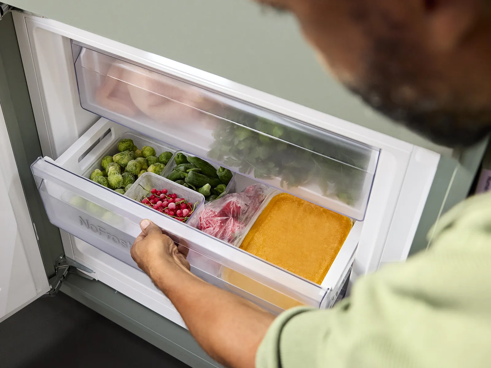 A person retrieves fresh vegetables and meats from a clear freezer drawer, highlighting organized food storage in a modern kitchen.