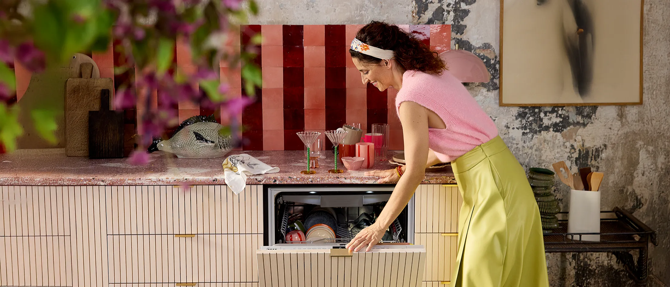 Sieviete rozā džemperī un zaļā svārkā līkumo, lai organizētu šķīvjus stilīgā virtuvē ar krāsainām flīzēm un dekoriem.