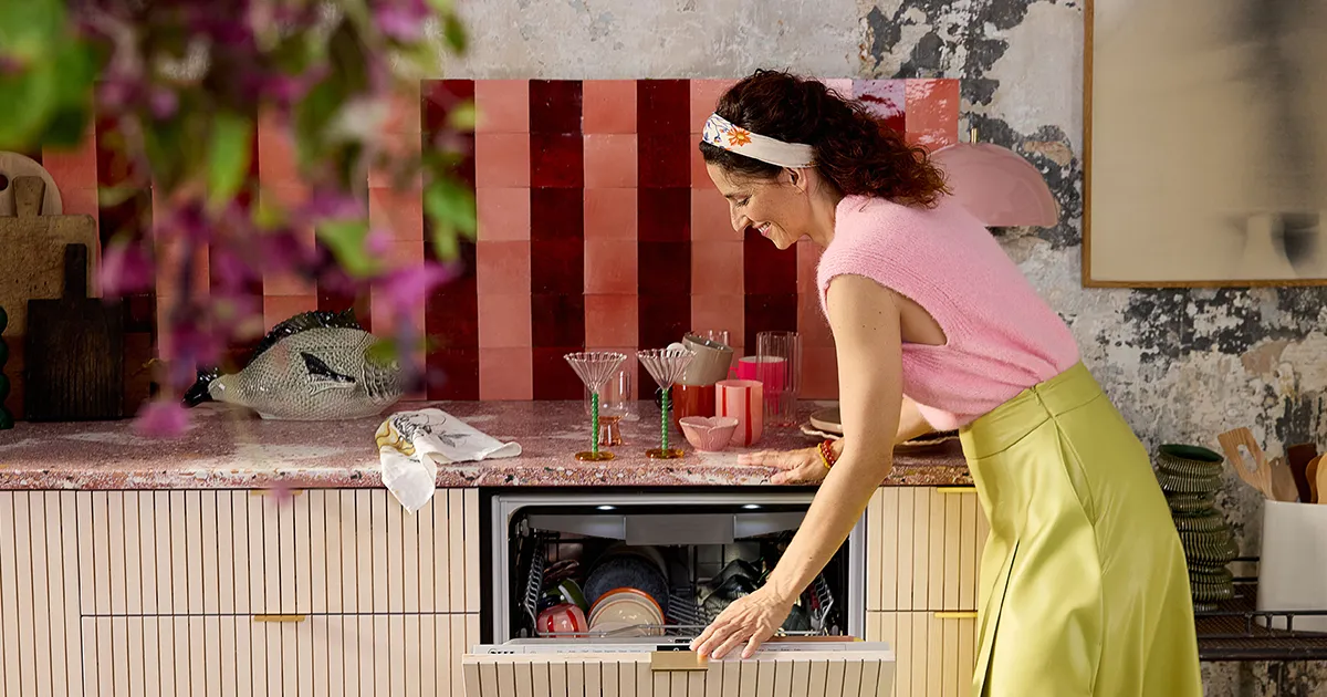 Sieviete rozā džemperī un zaļā svārkā līkumo, lai organizētu šķīvjus stilīgā virtuvē ar krāsainām flīzēm un dekoriem.