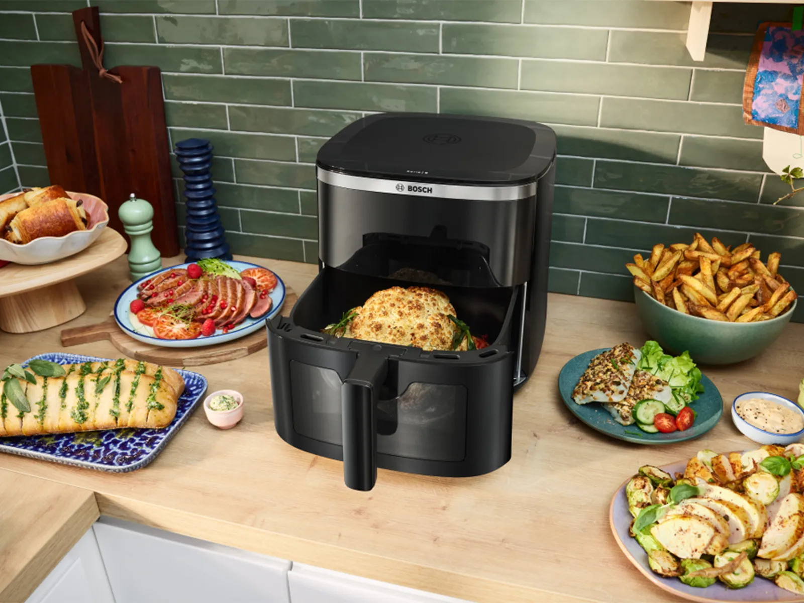 Φριτέζα αέρα Bosch σε πάγκο κουζίνας, περιτριγυρισμένη από διάφορα μαγειρεμένα πιάτα και συνοδευτικά.