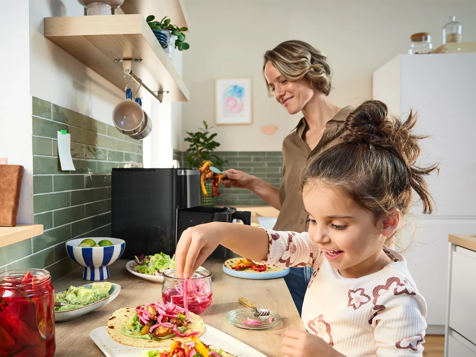 A woman and child, smiling, preparing food in a kitchen with fresh vegetables on the counter.