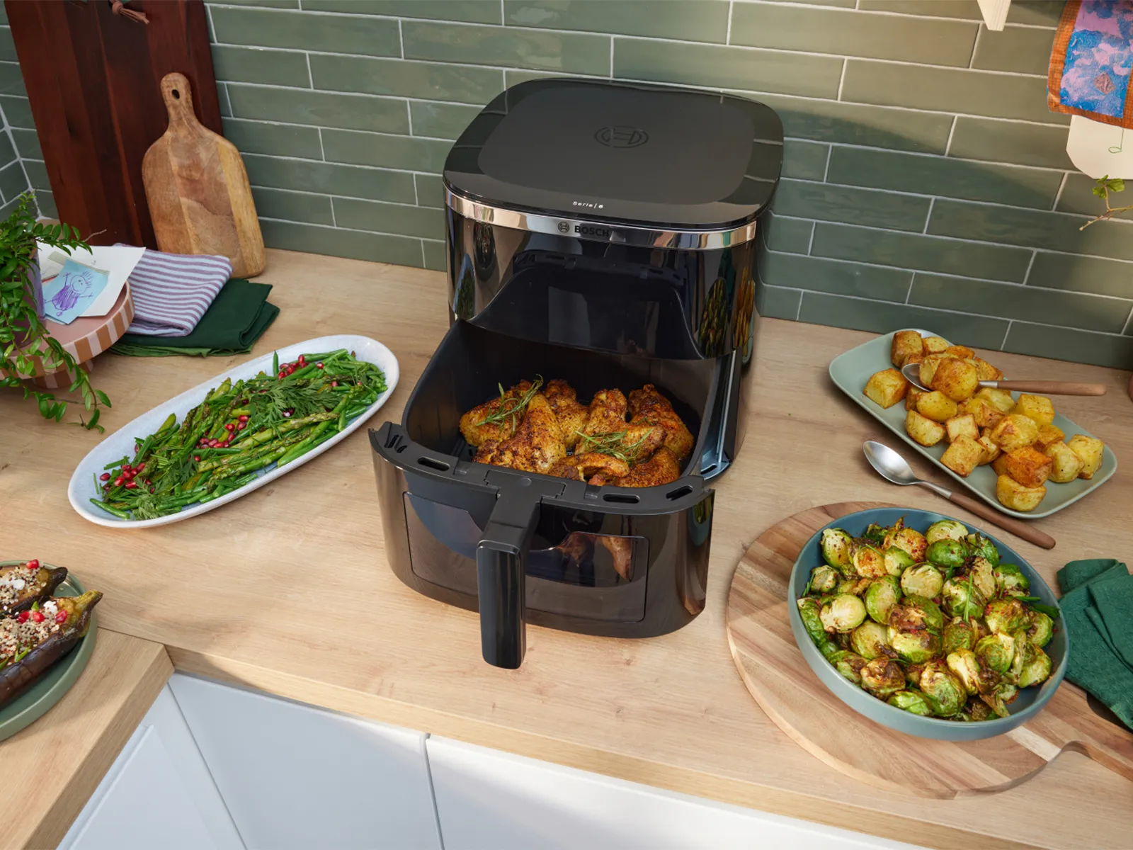 Bosch air fryer on a kitchen counter with food, including vegetables and chicken. Green tiles behind.