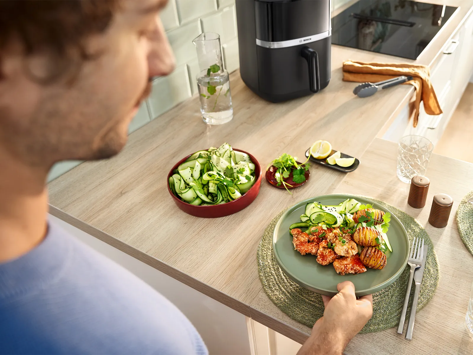 A young man places a plate of freshly cooked food on the kitchen counter. A second plate and a bowl of cucumber salad sit next to it.