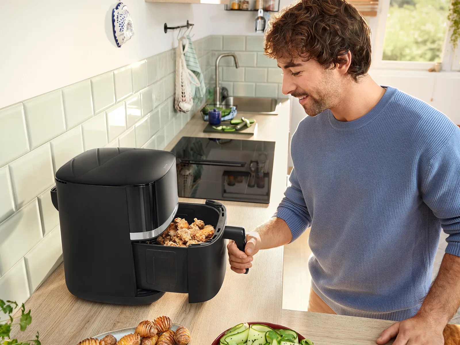 Gənc bir adam çiy yeməklə dolu bir qabı Bosch Hava Fritözünə sürüşdürür, parlaq bir mətbəxdə bişirməyə hazırlaşır.