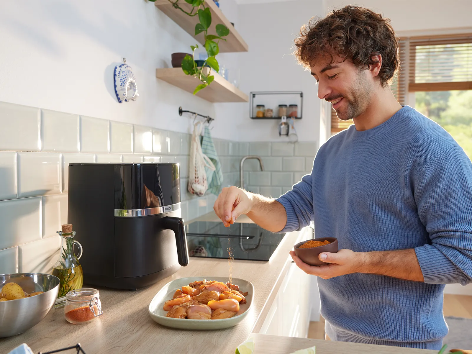 Tezgahda qara bir air fryer-in yanında mətbəxdə toyuğu ədviyyatlayan kişi.