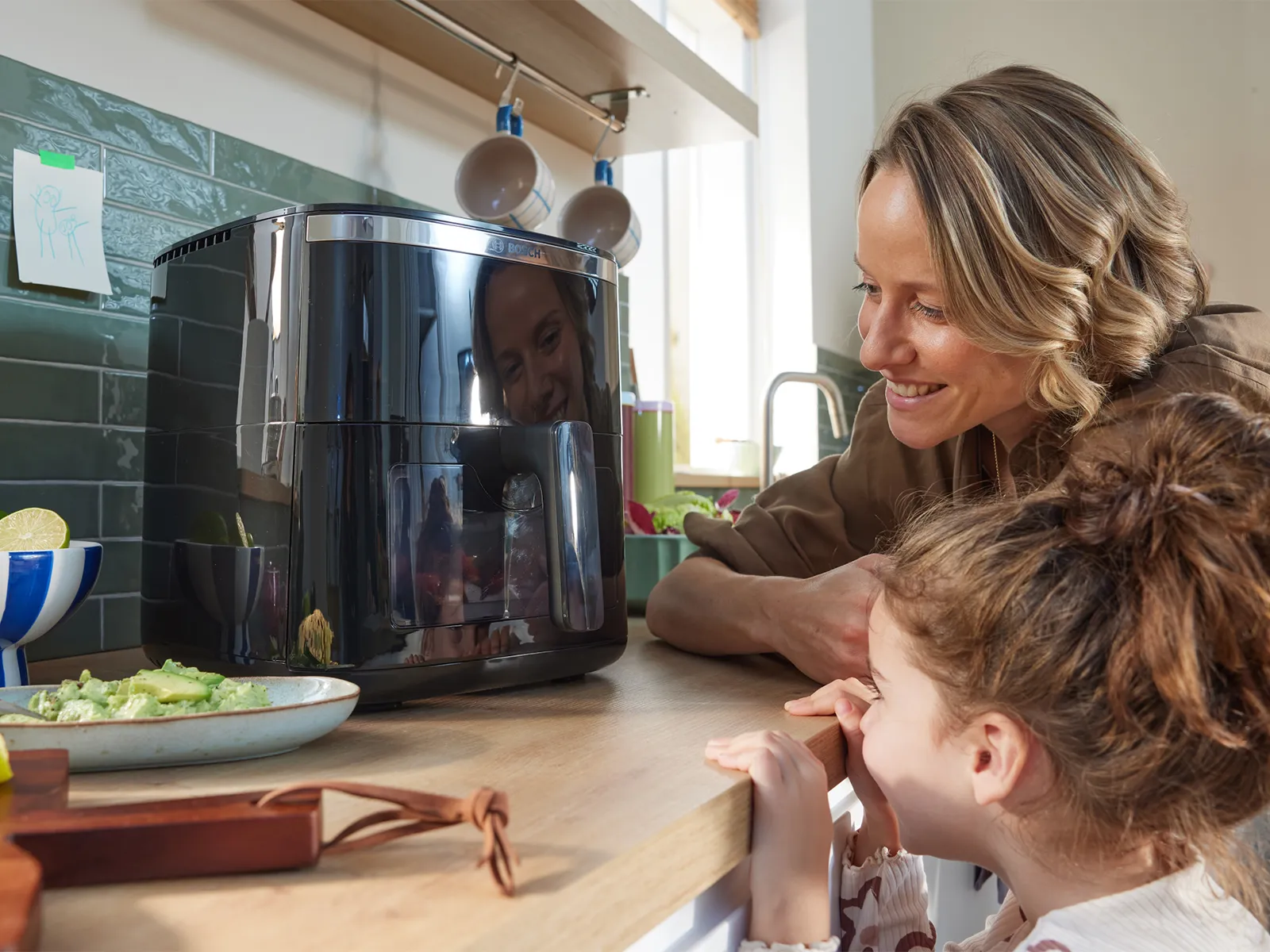 Ana və qızı yaxınlıqda yemək olan mətbəx tezgahında parlaq qara bir air fryer-ə gülümsəyir.