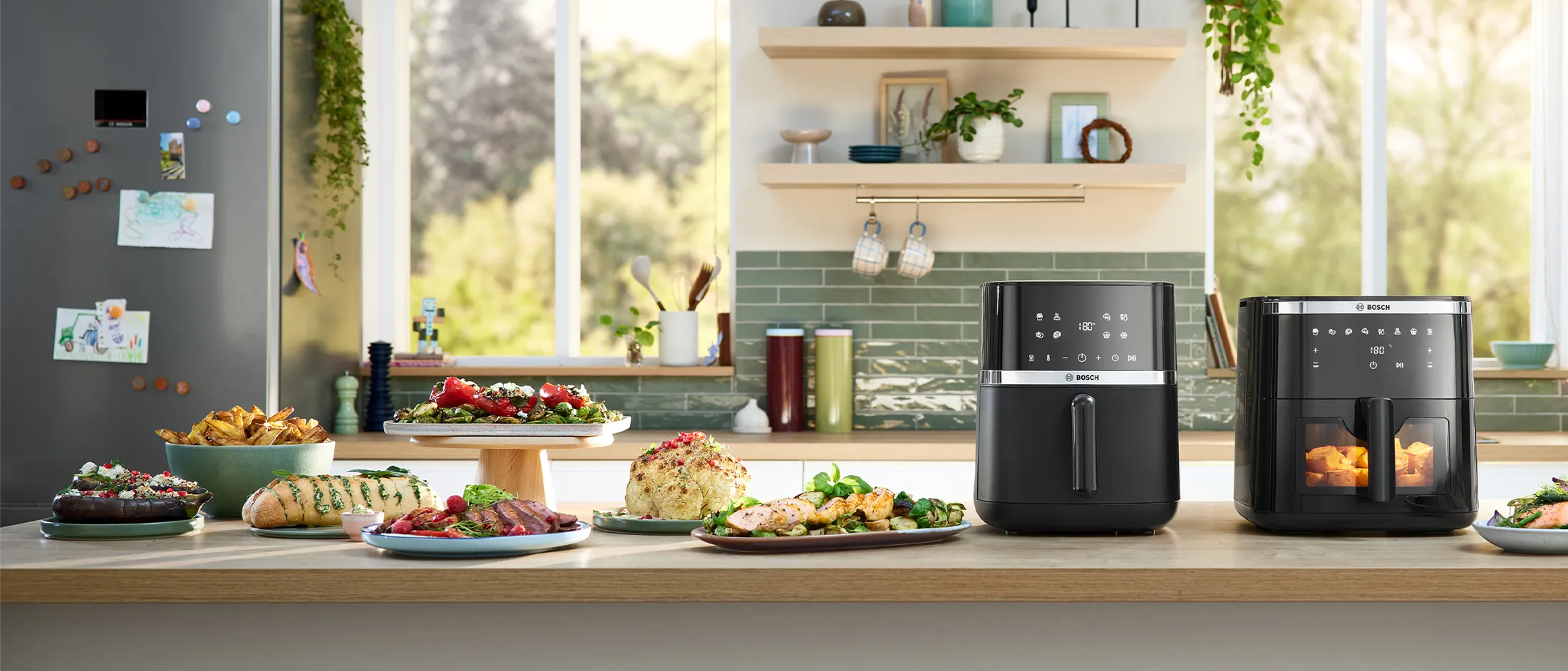 Dva modela Bosch Air Fryera jedan do drugog na svijetlom kuhinjskom pultu. Nekoliko tanjura i zdjelica s hranom, uključujući kruh sa začinskim biljem, piletinu, pečenje i krumpiriće, nalazi se pored uređaja i predstavljaju jela pripremljena pomoću Air Fryera.