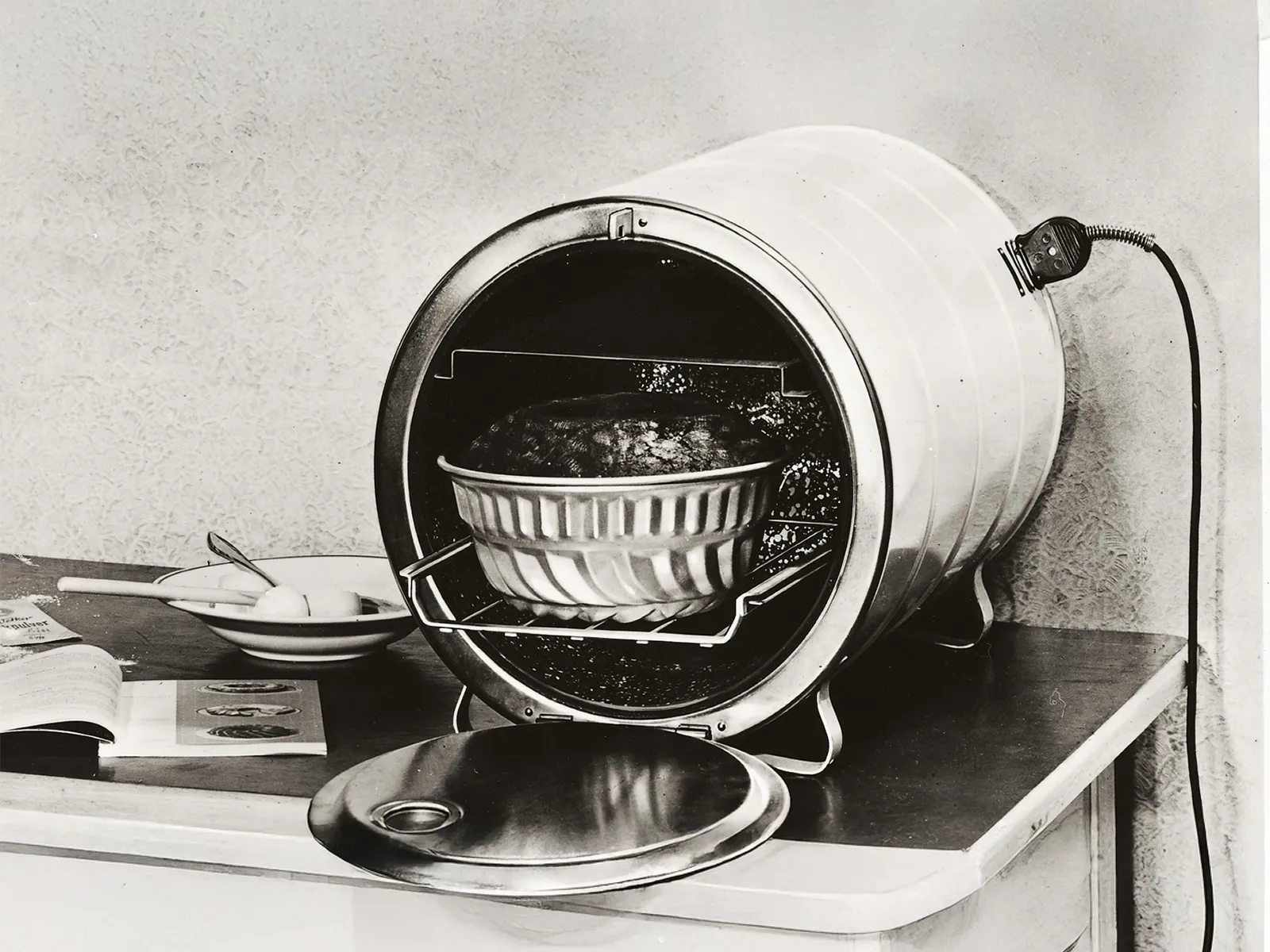 Forno elettrico vintage sul tavolo con teglia all'interno, coperchio aperto accanto, utensili da cucina e libro aperto sullo sfondo.