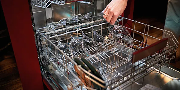 A hand placing a wine glass into the top rack of a dishwasher filled with clean glasses and utensils.