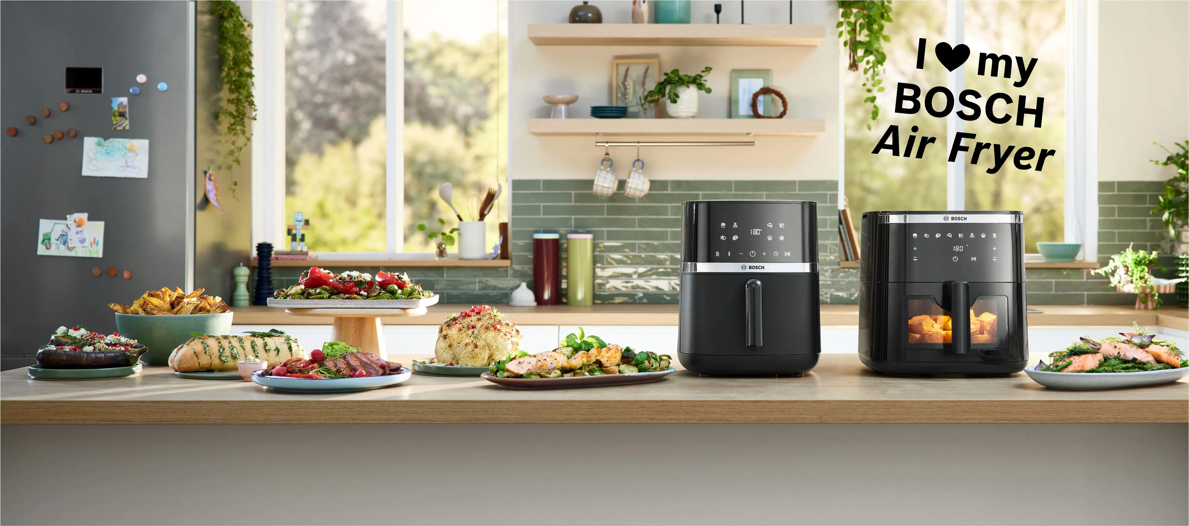 Două modele de friteuze cu aer cald Bosch aşezate alăturat pe un blat de bucătărie de culoare deschisă. Lângă aparate sunt aşezate mai multe farfurii şi castroane cu mâncare, inclusiv cu pâine cu mirodenii, carne de pui, friptură şi cartofi prăjiţi, etalând preparatele gătite în friteuzele cu aer cald, în coltul dreapta sus text "I love my Bosch Air Fryer"