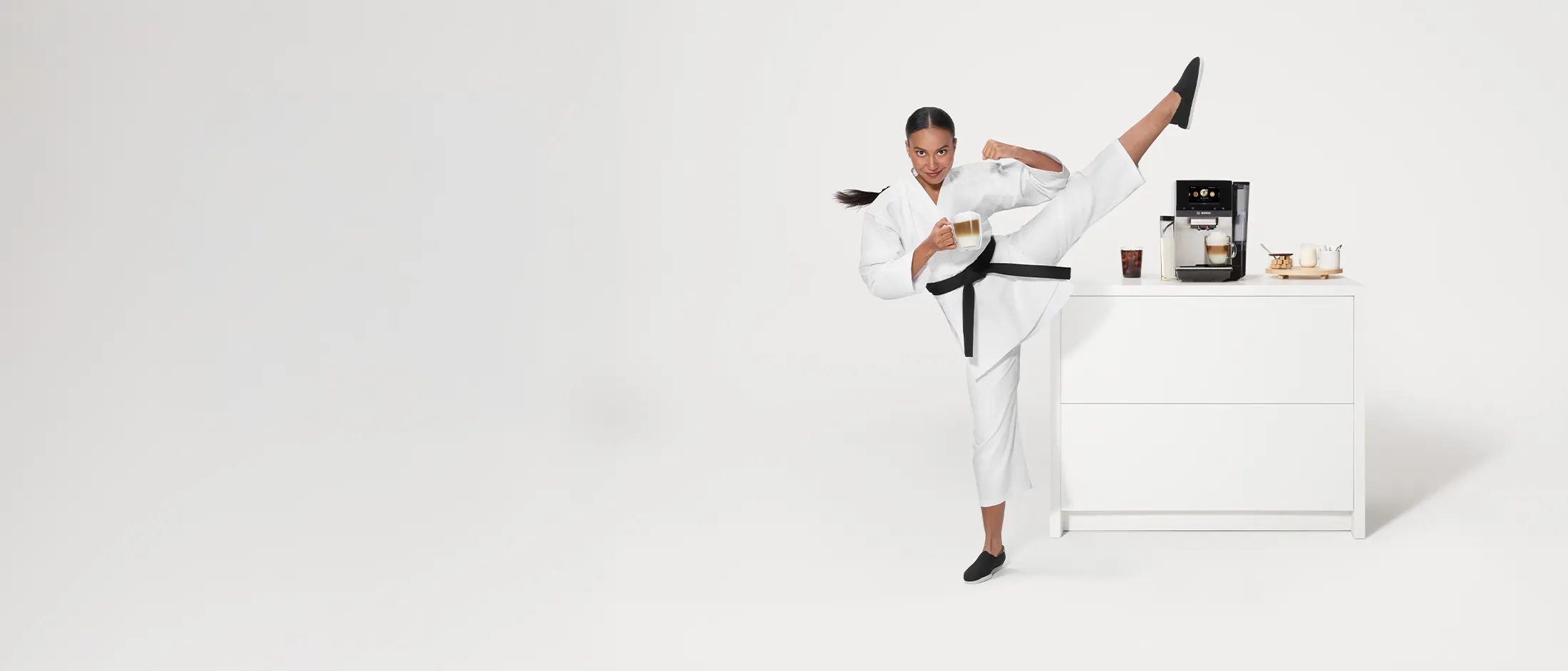 A person in a white karate uniform with a black belt performs a high kick in a bright, minimalist kitchen. Behind them is a sleek coffee machine and various cups, creating a dynamic blend of martial arts and culinary themes.