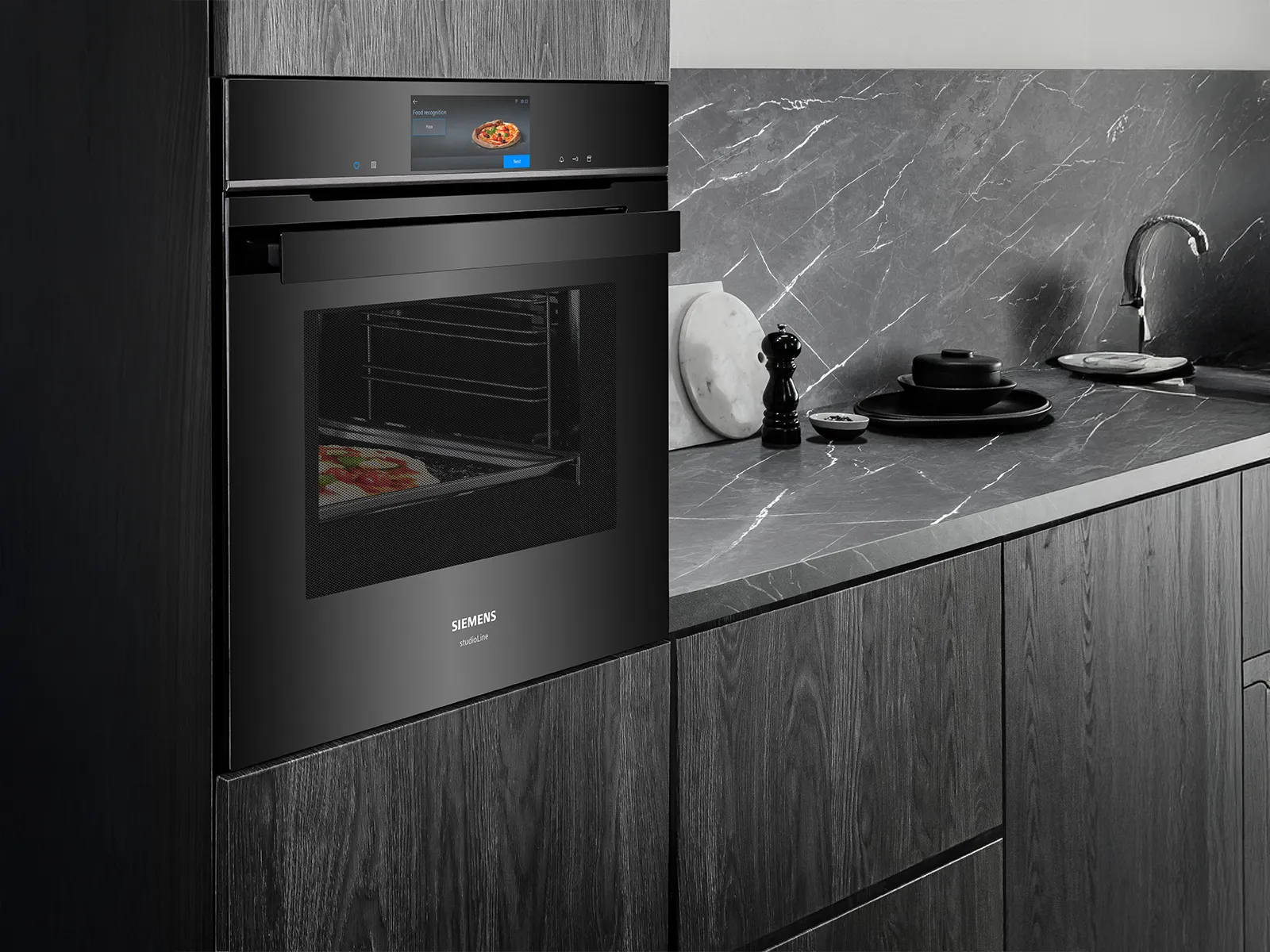 Un elegante forno Siemens nero integrato in mobili da cucina moderni, con un display digitale e una pizza all'interno, su un piano di lavoro in marmo.