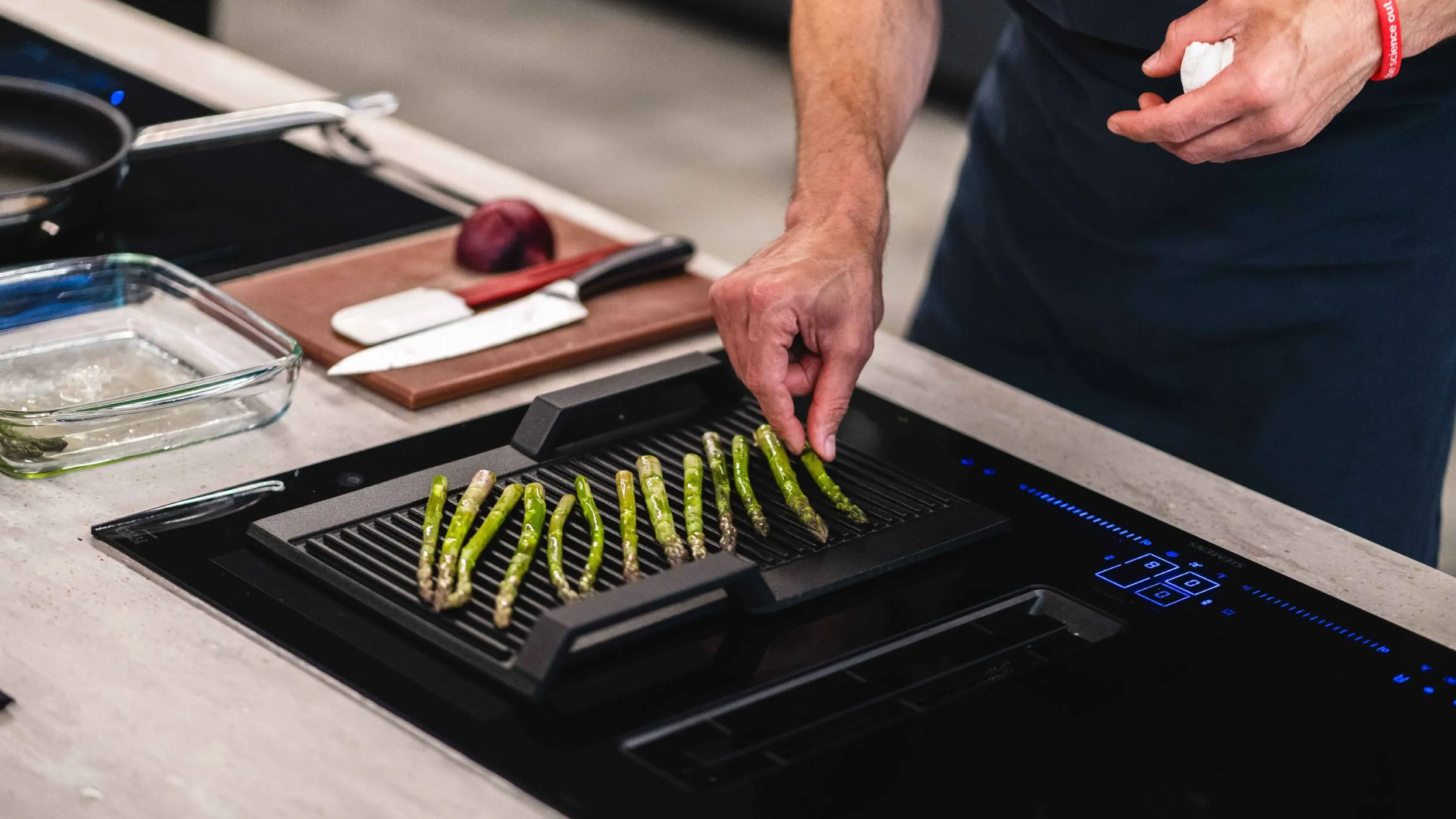 A chef is carefully arranging fresh asparagus spears on a grill pan placed on a sleek, black induction cooktop. In the background, a knife and a beet sit on a wooden cutting board.