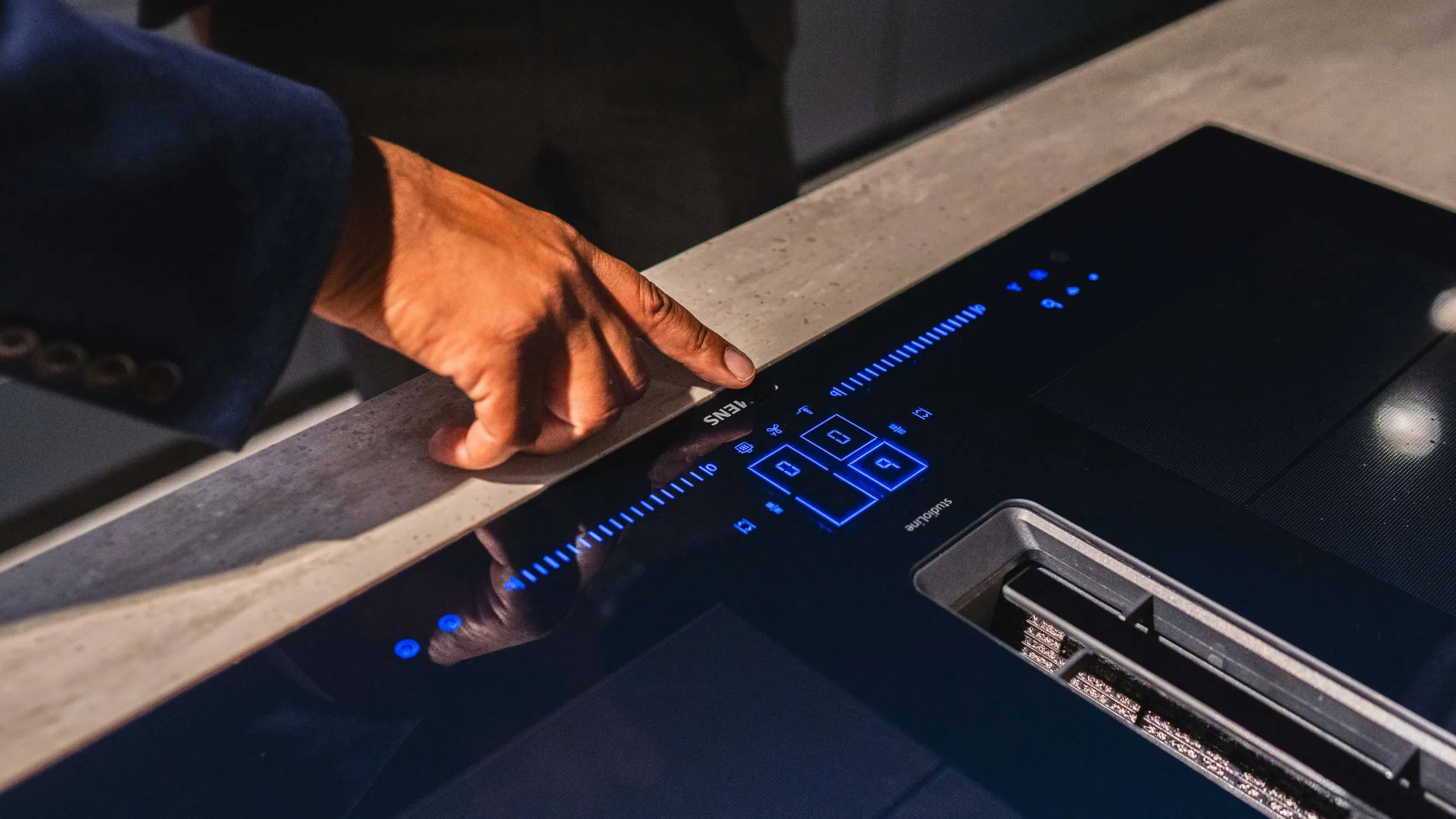 A person's hand is shown pressing the sleek, black touchscreen control panel of a modern induction cooktop. The cooktop is flush-mounted into a stone countertop and features illuminated blue digital displays and intuitive icons.
