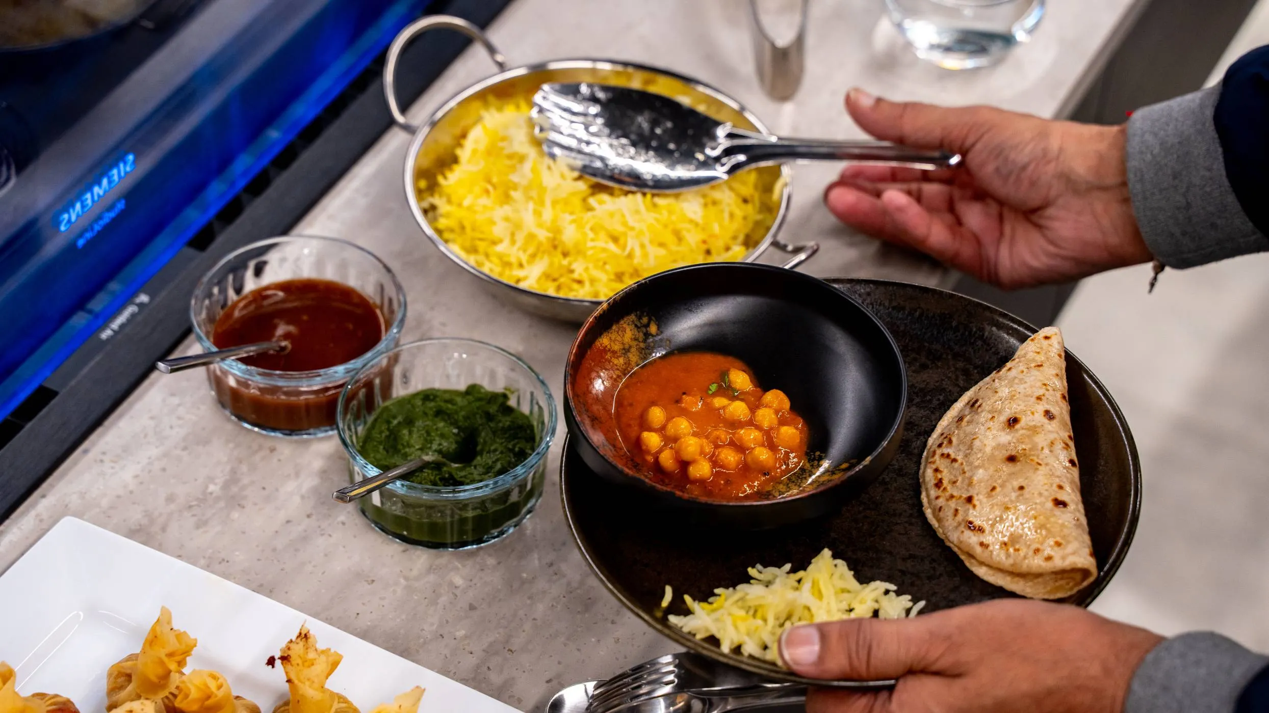 A person is plating a meal, holding a dark plate that contains a chickpea curry, a flatbread, and some grated cheese. In the background are bowls of rice, and green and red sauces.
