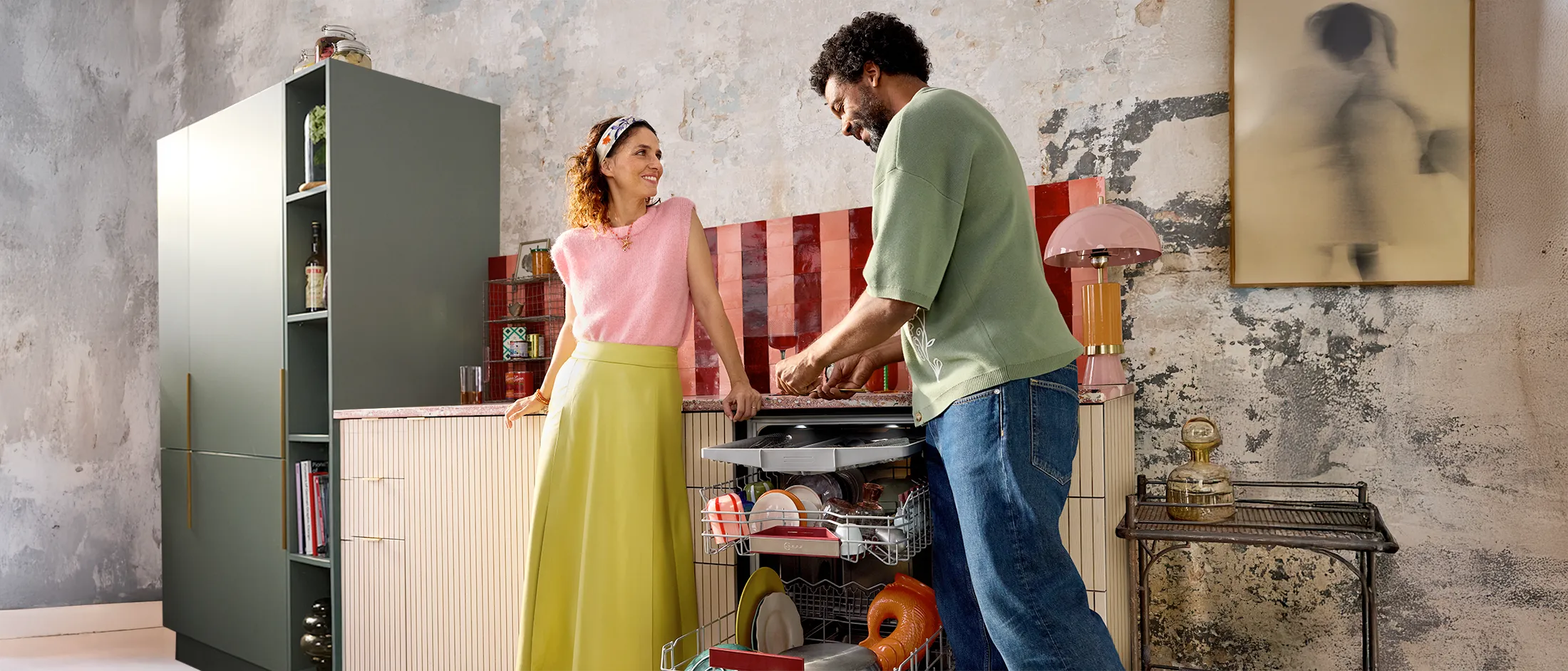En person i en grønn skjorte laster en oppvaskmaskin mens en annen person, kledd i en rosa topp og gul skjørt, ser på i et stilfullt kjøkken.