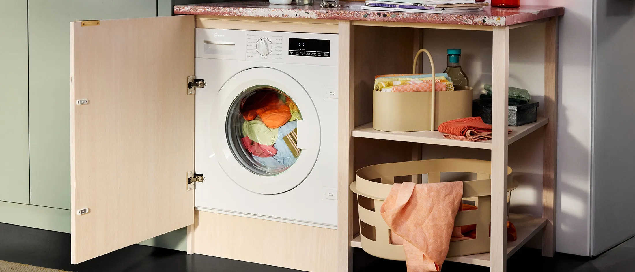 Waschbereich mit versteckter Waschmaschine, bunter Wäsche und organisiertem Regal mit Stoffen und Aufbewahrungsboxen.