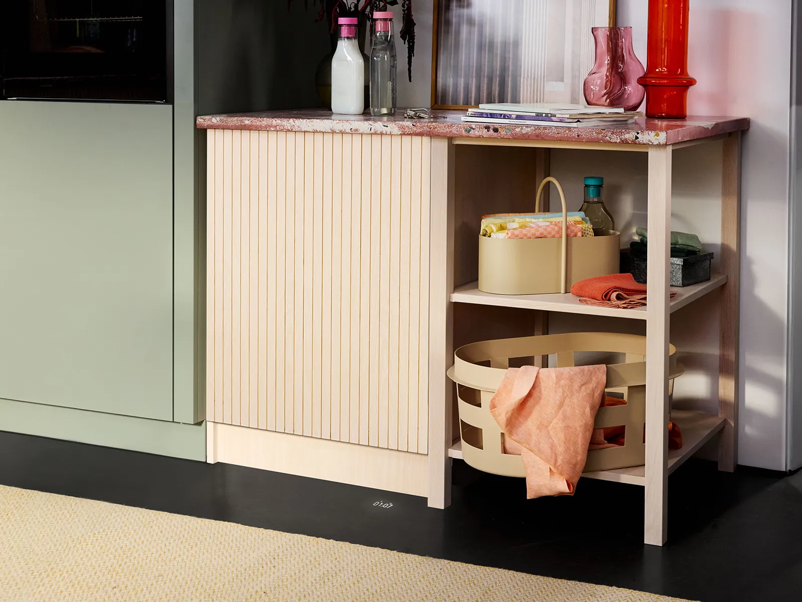 A stylish kitchen cabinet with a textured, light wood base and a pink marble top, featuring decorative items and colorful towels.