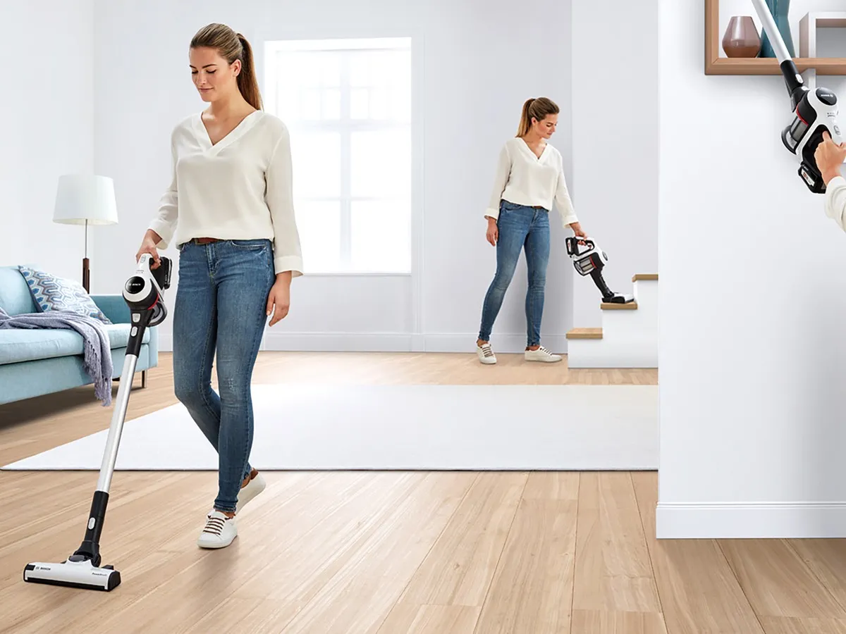 A woman vacuums the carpet in a tidy living room, surrounded by furniture and natural light from a window.