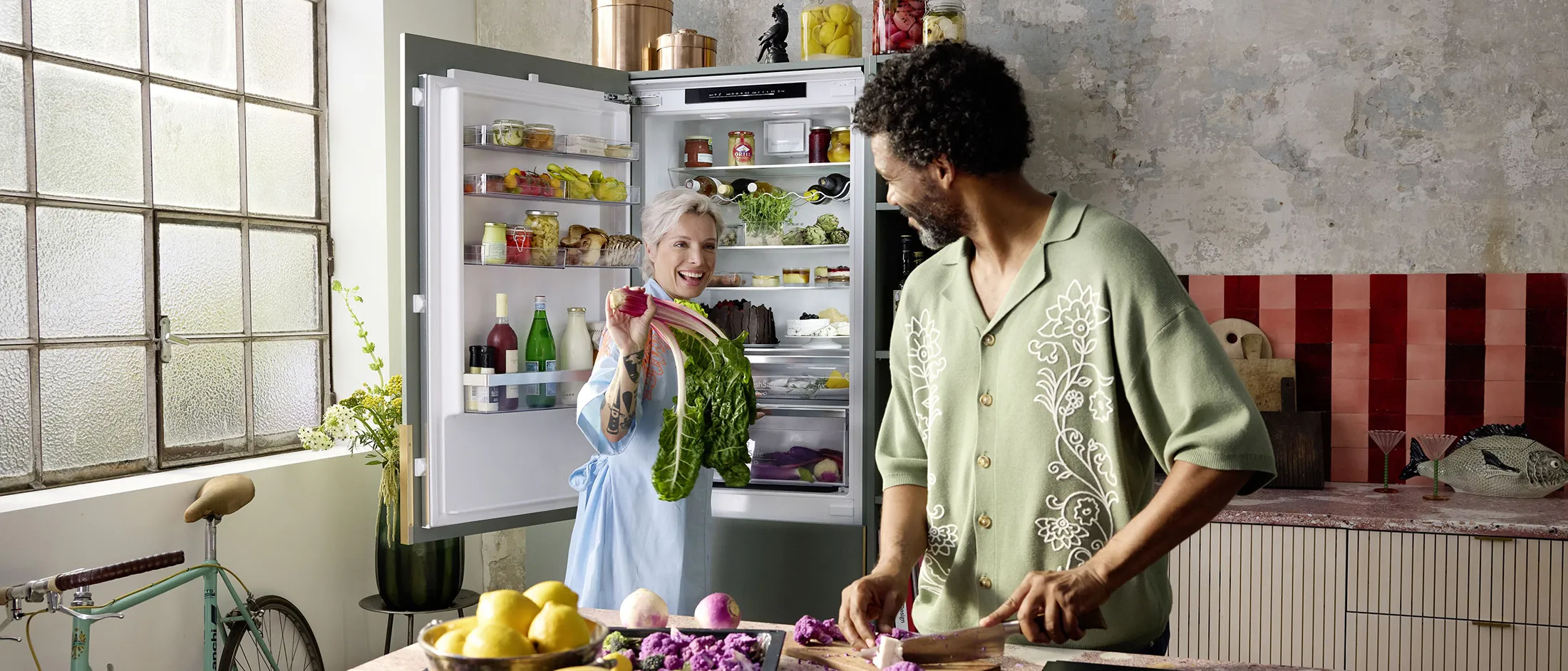 Isik valmistab toitu stiilses köögis, kus on avatud külmik, mis on täidetud erinevate toidukaupadega, ja taustal on jalgratas.