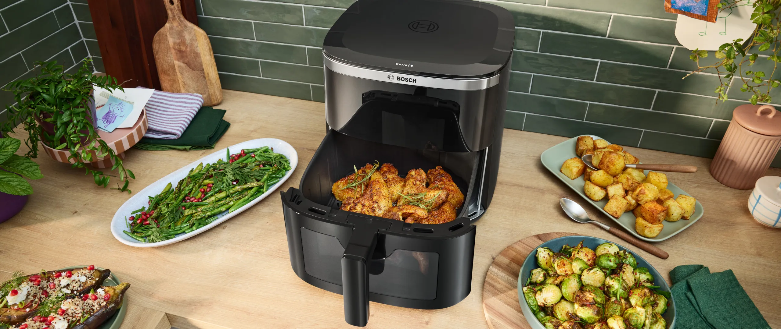 Bosch Air Fryer serije 6 s otvorenom ladicom za pečenje. Na obližnjem kuhinjskom pultu nalazi se zdjela prokulica, patlidžana i graha spremnih za pečenje.