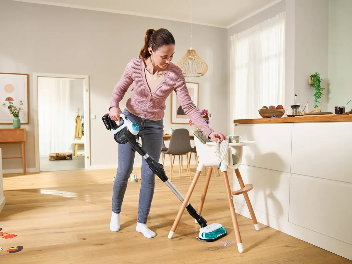 A woman vacuuming the kitchen floor, surrounded by cabinets and appliances.